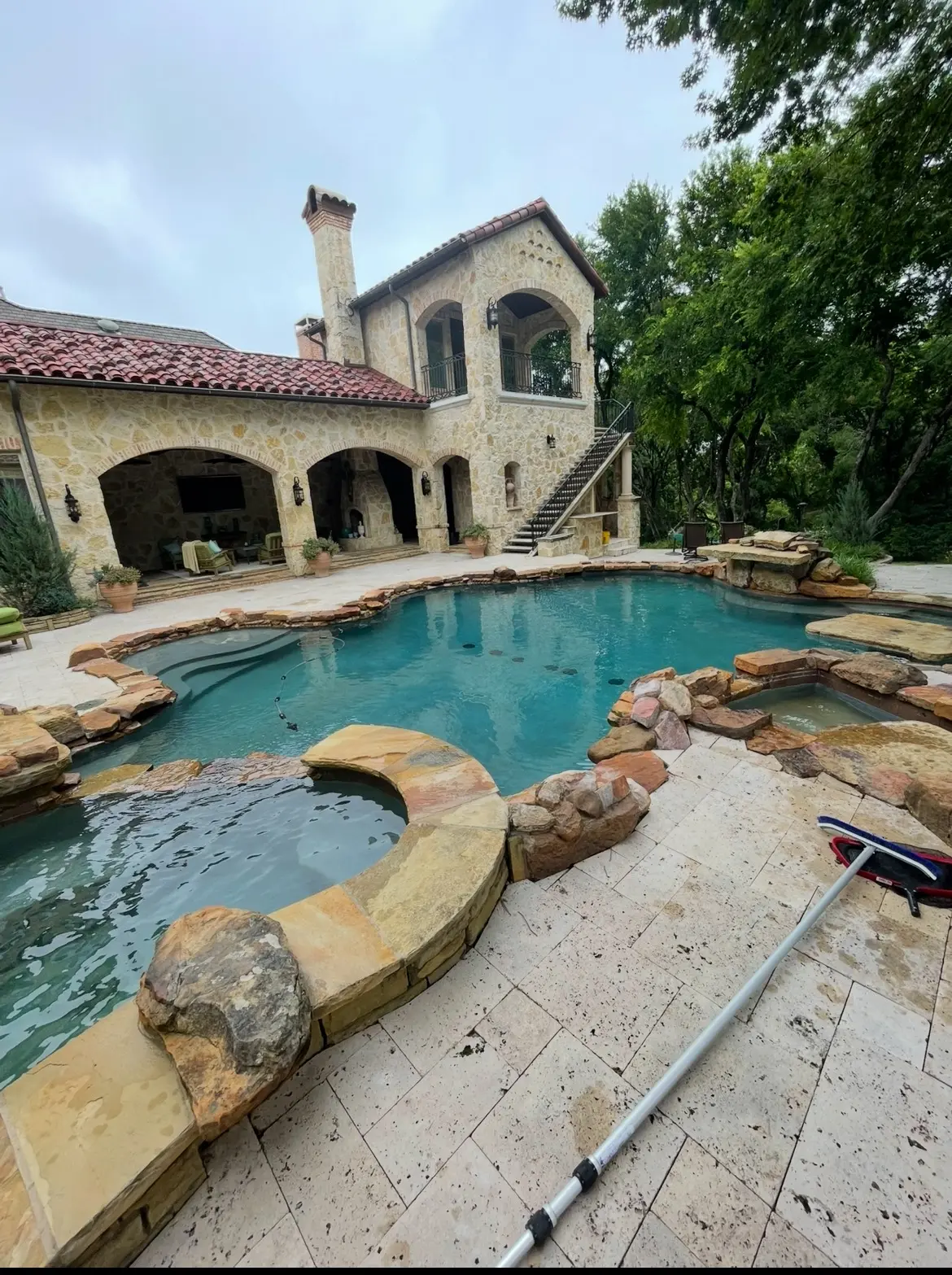 A stone Mediterranean style home behind a freeform pool with a raised rock spa and waterfall feature.